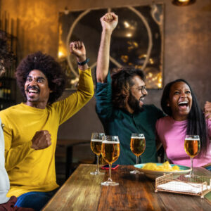 Multiracial people celebrating football team scoring goal in bar. Friends watching soccer game in pub. Leisure and sports concept.